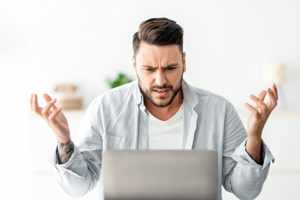 Angry millennial man looking at laptop screen and gesturing, having software, website error, sitting